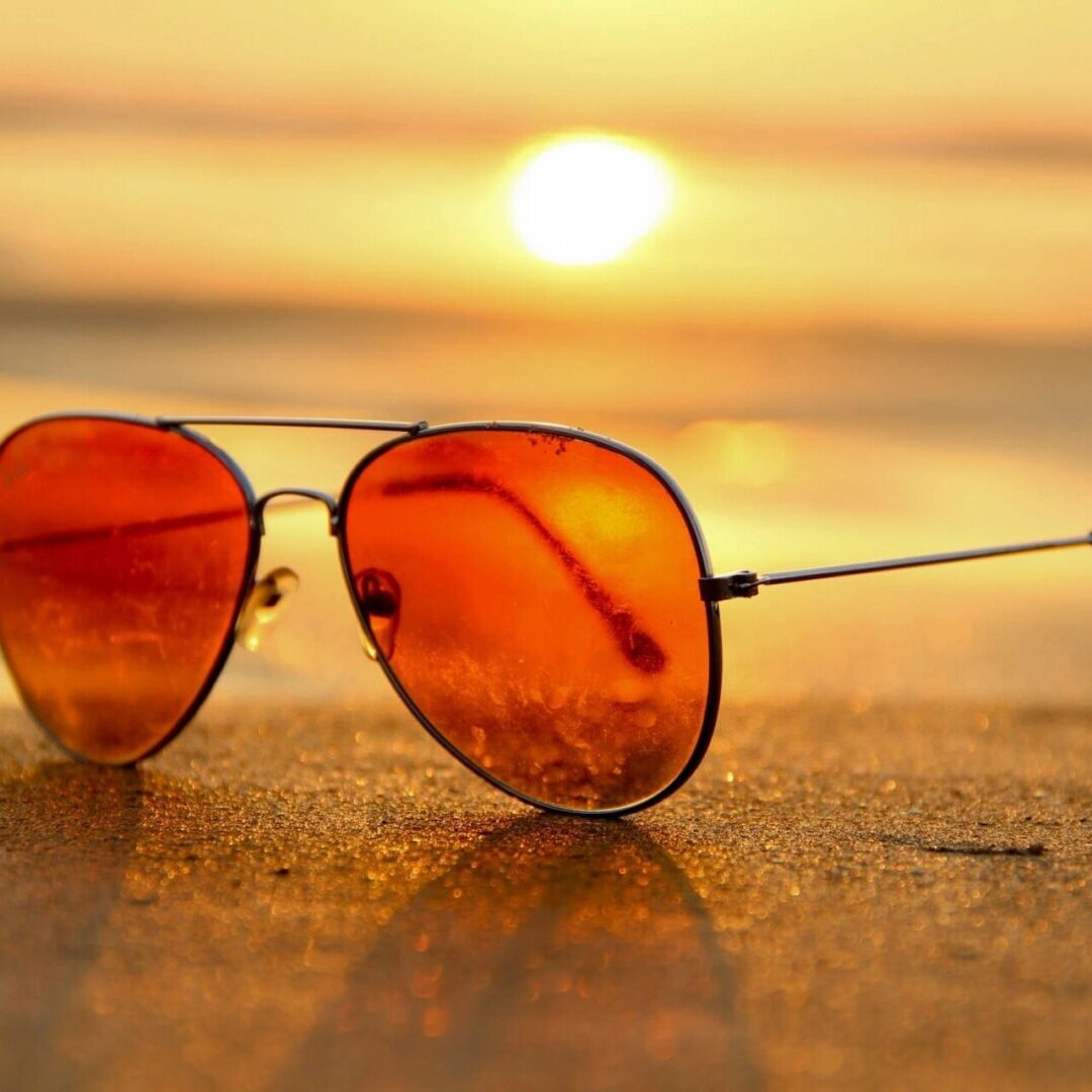 Orange-tinted sunglasses resting on sand with the sun setting in the background.
